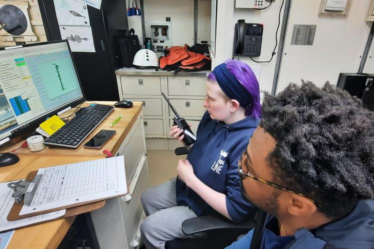 Two people at a desk in front of a computer and monitor in a dry lab on a research vessel. The computer displays a dark green vertical line. Kenzi Kimball is in the center of the image, with purple hair bound in a bandanna and wearing a blue hoodie, holding a walkie-talkie to give instructions to a winch operator out on deck. Audy Peoples sits nearest the camera, watching the screen.
