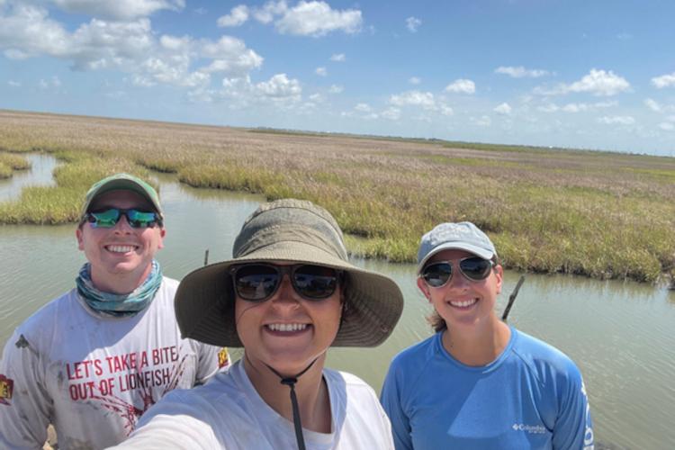 Three people in hats and sunglasses stand smiling in a marsh.