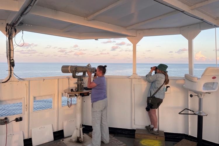 2 women stand on the deck of a ship looking through binoculars