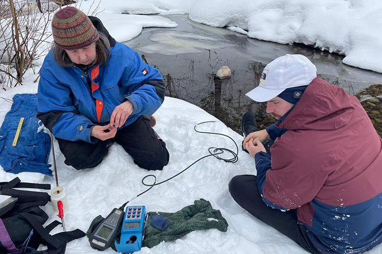 Two people sit on the snow along a creek. They are both looking at a piece of scientific equipment. One is wearing a blue winter jacket and knitted beanie, and the other is wearing a red and blue winter jacket and white baseball cap.