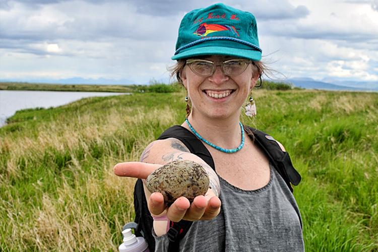 Woman with green hat holding a rock with grass and water in background