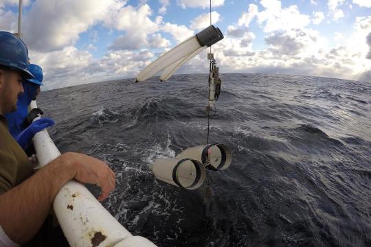 Two men on deck at the rail of a ship watching a fine-mesh net array being hauled onboard by a winch.