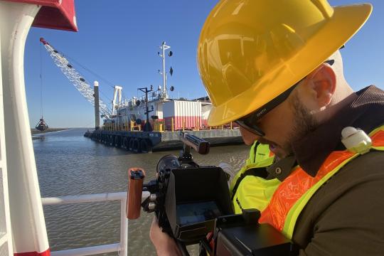 A man wearing a safety vest and hard hat takes a photograph of a barge