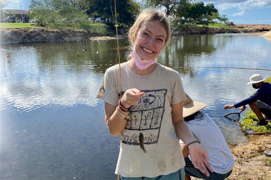 Hollings scholar is holding a fishing pool with a caught fish on the line near a pond.