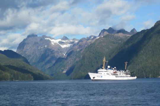 A large white ship with the NOAA insignia floats in a bay surrounded by mountains.