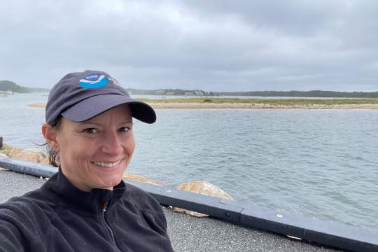 Woman scientist wearing NOAA hat and smiling stands in front of blue water with land and marsh grass in the distance.