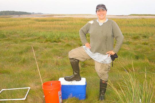 Genevieve wears a tan ballcap, dirty clothes, and field boots in a marsh. Her right foot is on top of a cooler and her hands are on her hips.
