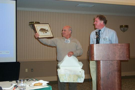 One man holds up a framed fish print presented as a thank you gift while a second man stands to his right behind a podium.