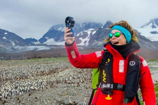Catherine Foley wears a fleece ear and head band, sunglasses, orange winter foul weather jacket and a personal floatation device while holding a wind measuring instrument in her right hand. Behind her is a large colony of penguins. There are snow-covered mountains in the background and the sky is clouded over. Some of the clouds are low and hide the mountain tops.