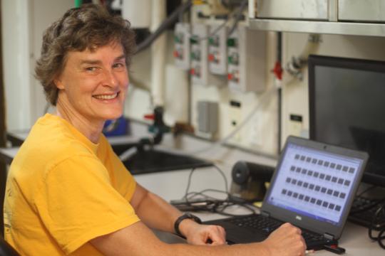 A woman in a yellow t-shirt with short, curly hair sits in front of a laptop and smiles at the camera.