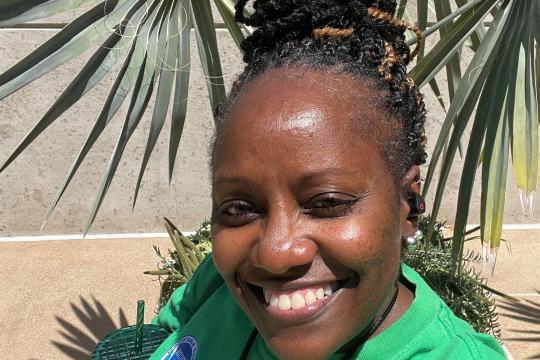 A woman in a green t-shirt smiles for the camera while standing in front of a concrete wall and leafy plants.