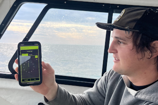 A man in a gray hoodie and hat holds up his phone to the camera while sitting inside a ship cabin.