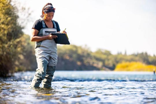 Dereka Chargualaf wading in a river, writing on a clipboard