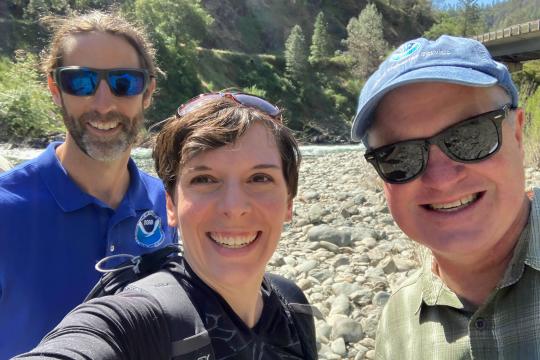 Three people smiling for the camera, standing on a rocky riverbed