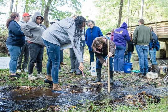 Two women lean over to examine how one of the women is holding a yardstick in a stream as other people look on. Credit: Virginia Commonwealth University.