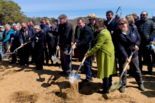 Carrie Selberg Robinson (front row, right) stands in a crowd holding a shovel at groundbreaking ceremony for Herring River Restoration Project in Cape Cod, MA. 