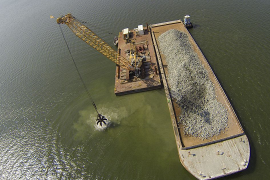 An aerial view of a crane moving hard substrate from the deck of a barge into the Piankatank River
