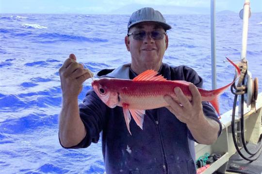 A fisher smiles while holding up a red and white fish, called an onaga, with both hands while out at sea.