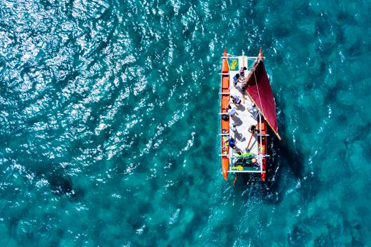 A bird's-eye-view of eleven people on a canoe over clear blue waters.