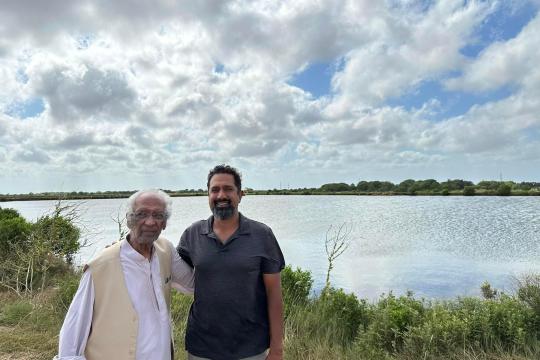 An adult man and his father stand with arms around each other with saltmarsh and a fish pond in the background.
