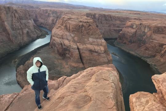A woman in jeans and a jacket standing near the edge of a massive horseshoe shaped canyon with water at the bottom.