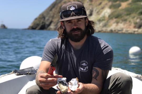 A bearded man wearing a hat on a boat holds a freshly shucked oyster.