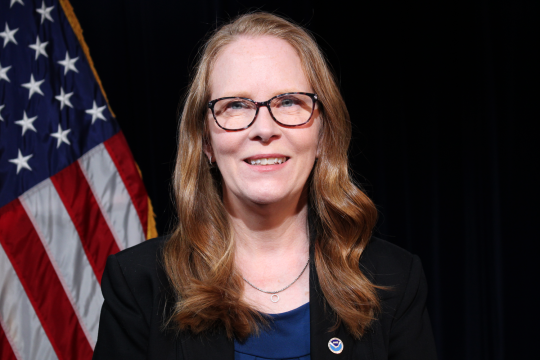 A red-haired white woman wearing glasses and a blue shirt with a black blazer sits in front of an American flag and a black background.