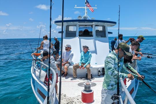 Roughly one dozen people, many wearing hats and active wear, stand along the railings of a white fishing boat, holding fishing poles.