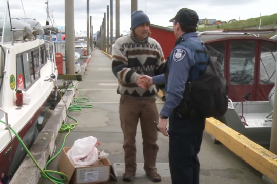 Image of a man in a sweater and a man in uniform shaking hands on a dock surrounded by boats