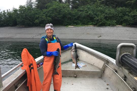 Teacher at Sea and Commercial Fisherwoman, Germaine Thomas, stands at the bow of her boat. She is dressed in orange coveralls, cold weather gear, and is holding two salmon--one in each hand.