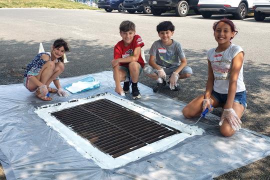 Four young students kneel by a storm drain. They are working on painting the drain with information about the Chesapeake Bay.