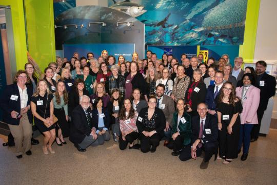 A large group of people smile in front of an ocean-themed backdrop in a museum