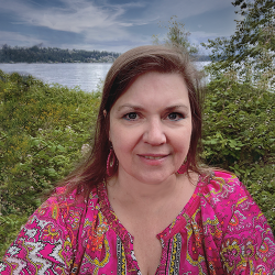 Rebecca white stands in front of blackberry brambles with Lake Pend Orielle in the background.