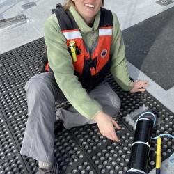 Lindsey Peavey Reeves sits on the back deck of the R/V Fulmar next to a SoundTrap underwater recorder. She looks up and is smiling at the camera.