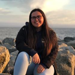 Kourtney Burger sitting on the rocks with the ocean in the background. This is her bio photo and she is facing the camera and smiling