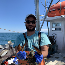 Isaiah holding lobster on fishing vessel, wearing blue t-shirt and raingear.