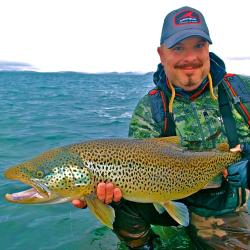 Man in blue hat and waders stands in waist deep water holding a large green/yellow fish with brown spots.