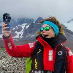 Catherine Fley, wearing red jacket and blue headband with anemometer.  Mountains and penguins in the background.