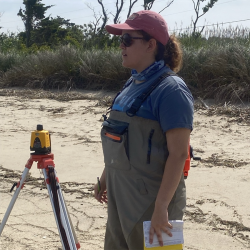 A young woman wearing sunglasses, a ball cap, and hip waders stands on a beach next to survey equipment