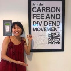 A smiling young woman points to a sign that reads "Join the Carbon Fee and Dividend Movement"