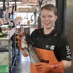 Blonde, short-haired woman smiles and poses with long, narrow silver and black fish she's holding while wearing orange gloves and bib pants.