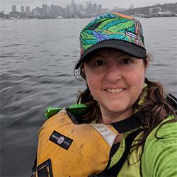 Scientist wearing vest and hat on boat with water and city in background