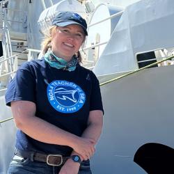 Blonde woman poses in NOAA Teacher at Sea shirt and hat stands and smiles in front of a white ship.