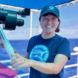 An asian woman wearing a blue NOAA Teacher at Sea cap and shirt smiles for the camera as she holds onto two metal handles on a ship