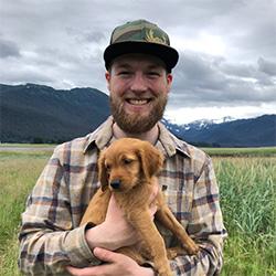 Portrait of scientist wearing a hat and holding a puppy with a field and mountain in background