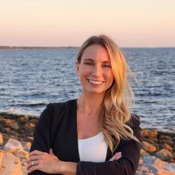A woman with long blonde hair smiling with the ocean behind her