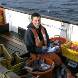 dam-migration-miss/md_0Q8MgoctYdzh.jpg Male fishery observer kneels on a boat with tubs of fish while holding a clipboard and pen and smiles for camera.