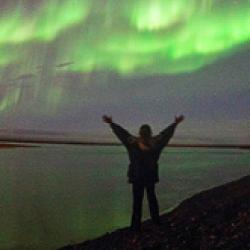Woman with arms extended near the water with green lights in the sky 