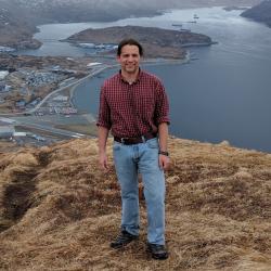 Brunette man with red shirt and jeans on a hill overlooking a town, water and hills