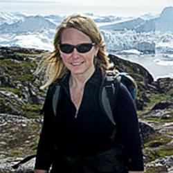 Woman wearing glasses with black sweater and backpack with mountains in background. 